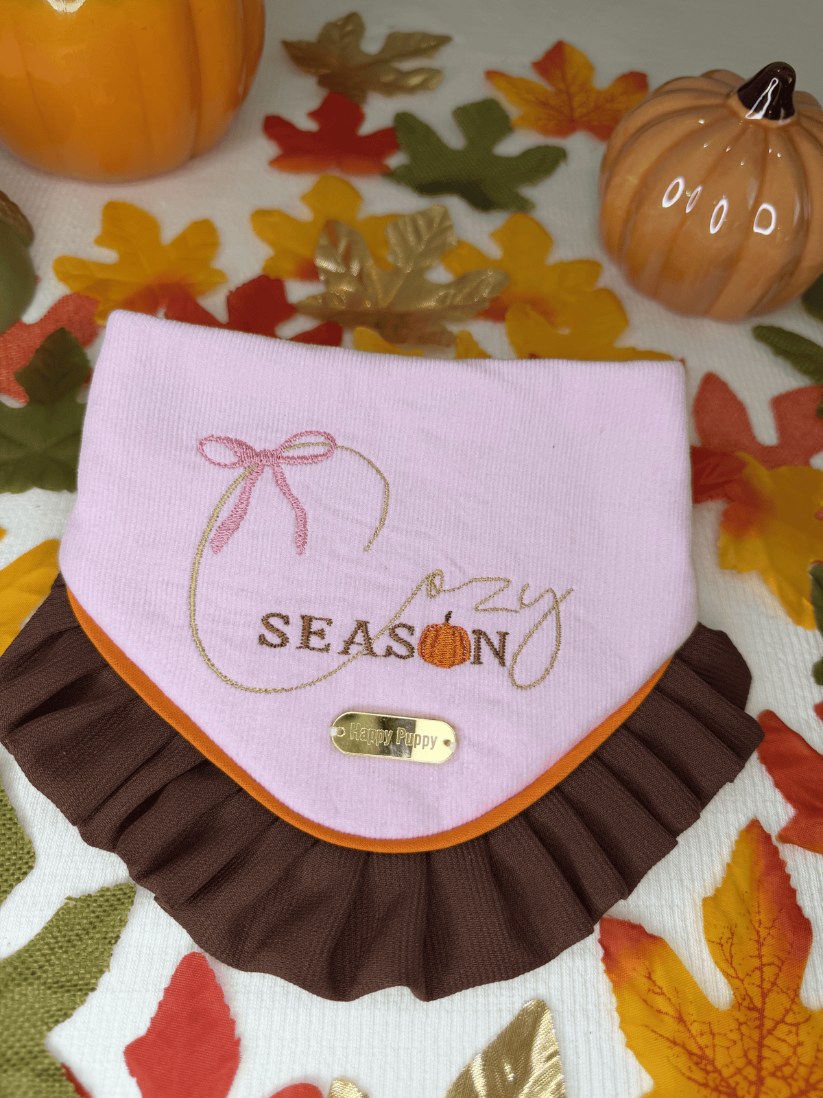 Bandana para perro Cozy Season de Happy Puppy Bandanas en rosa con las palabras Cozy Season y un lazo bordados, ribete naranja y volante marron, vista frontal