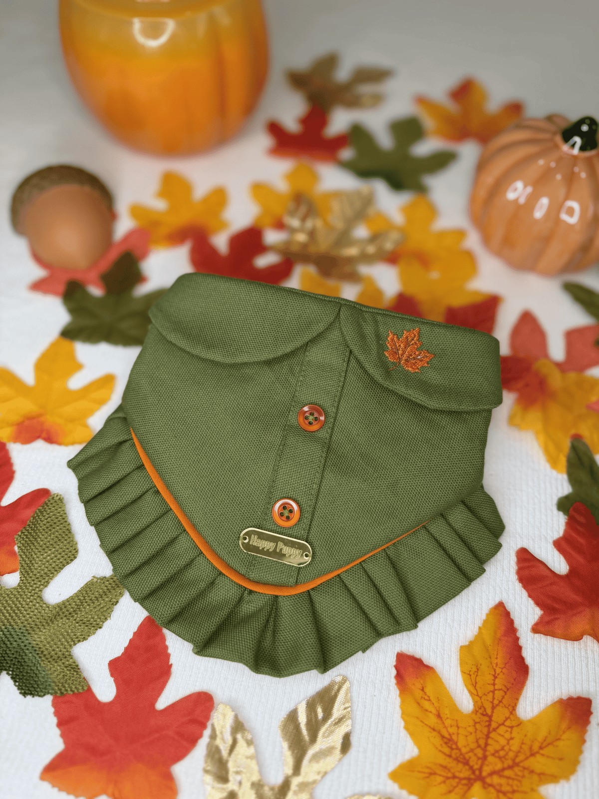 Detalle de la bandana para perro Pumpkin Patch de Happy Puppy Bandanas en verde oliva con botones y volante naranja, sobre hojas de otono