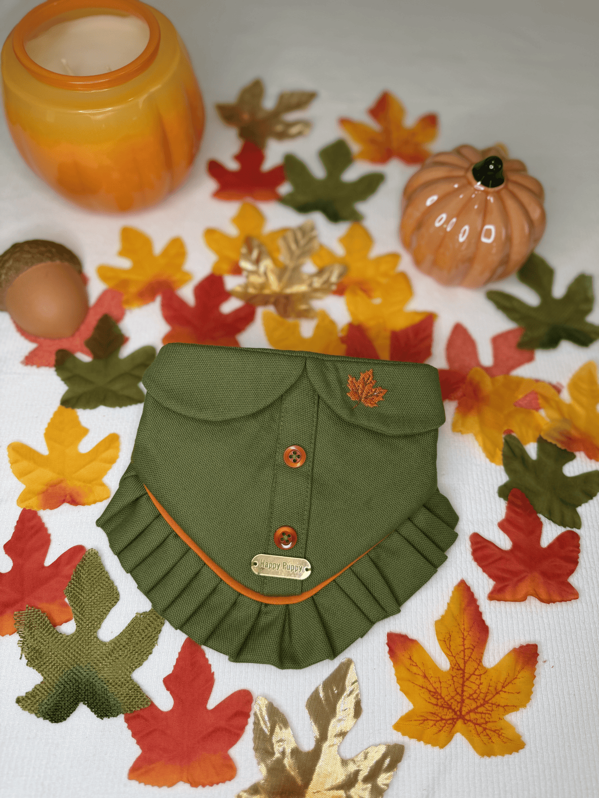 Bandana para perro Pumpkin Patch de Happy Puppy Bandanas en verde oliva con botones naranja y volante naranja, vista frontal sobre hojas y calabazas de otono