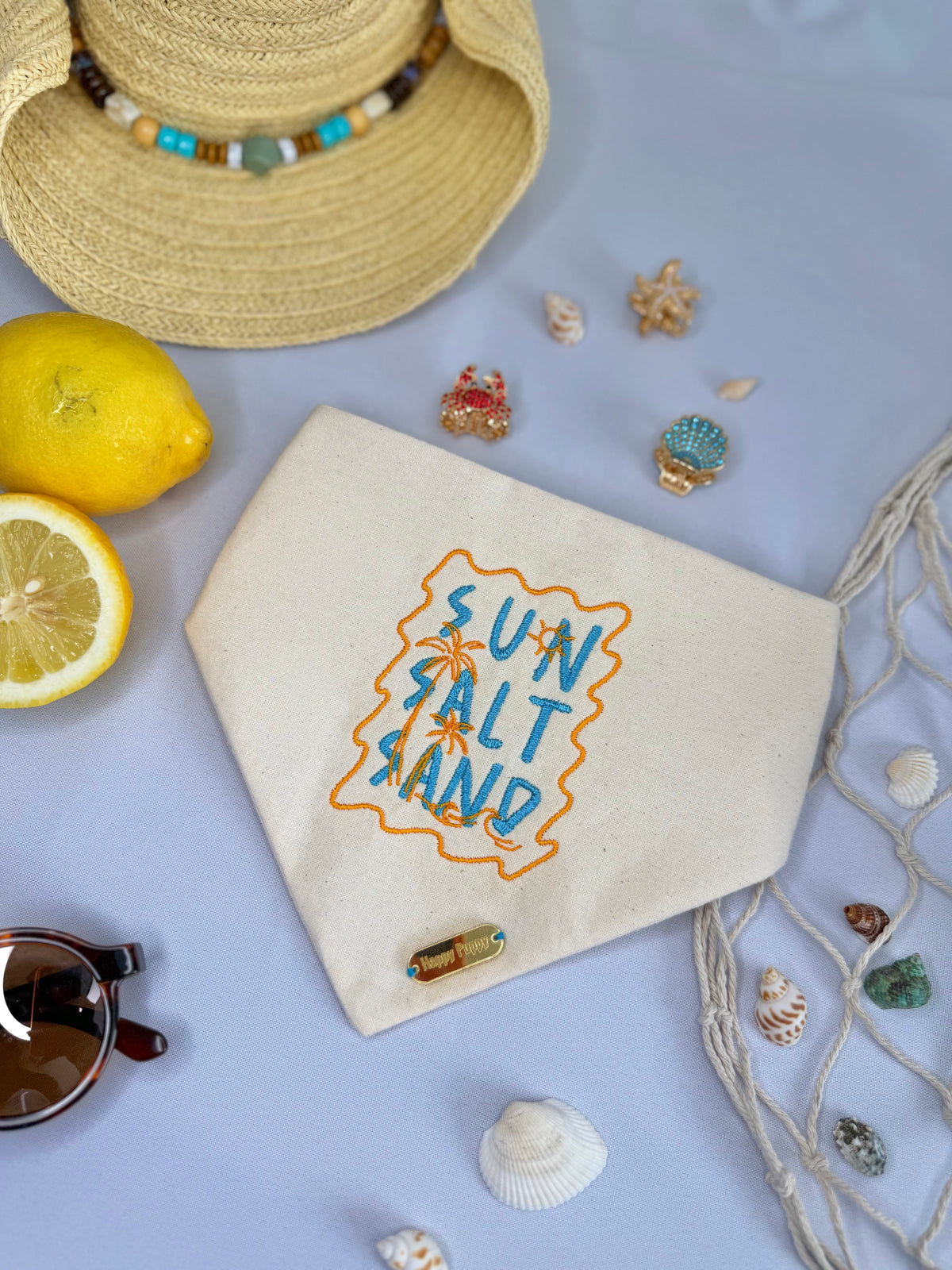 Bandana para perro Sand de Happy Puppy Bandanas en crema con las palabras Sun Salt Sand bordadas en azul y naranja, vista frontal en ambiente de playa